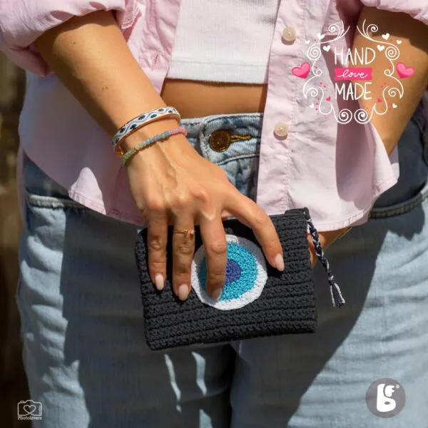 Χειροποίητο πλεκτό mini wallet σε μαύρο χρώμα με σχέδιο μπλε ματιού και φερμουάρ, κρατημένο στο χέρι σε καθημερινό καλοκαιρινό outfit.